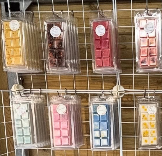 A retail display of assorted wax melts and candles arranged on a white wire grid against a wooden wall. The display includes many small packaged wax melts in assorted colors, several black jar candle containers, amber glass candle jars, and a blue digital scale on a shelf.